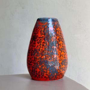 Op de afbeelding: Een druppelvormige keramische vaas met een levendig oranje gespikkeld patroon en een donkerblauwe rand. De vaas heeft een glanzende afwerking en een unieke, getextureerde uitstraling. Deze decoratieve vaas is geschikt voor interieurdecoratie.