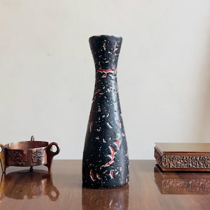 Puede incluir: Un jarrón negro mate, alto y esbelto, con la parte superior acampanada y una superficie texturizada que presenta motas irregulares rojas y blancas. El jarrón tiene un cuerpo cilíndrico cónico que se ensancha hacia la base y se estrecha en el cuello.