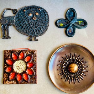 May include: A collection of vintage metal wall art pieces. Includes a lion, a four-petal flower, a square tile with a flower design, and a round plate with a sunburst pattern. The pieces feature bronze, copper, and enamel finishes.