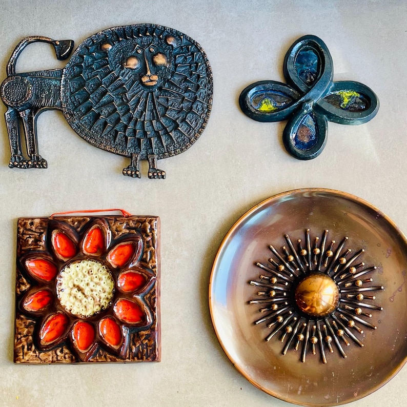 May include: A collection of vintage metal wall art pieces. Includes a lion, a four-petal flower, a square tile with a flower design, and a round plate with a sunburst pattern. The pieces feature bronze, copper, and enamel finishes.
