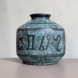 May include: A teal ceramic vase with a textured surface and a dark brown rim. The vase features embossed zodiac symbols around its body. The design is a mix of teal and brown, creating a vintage aesthetic. The vase is a decorative item.