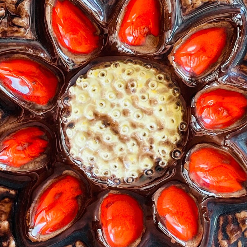 May include: Close-up of a decorative ceramic piece with a central yellow and brown circular design surrounded by bright red, teardrop-shaped elements. The piece has a glossy finish and is set against a dark brown background, creating a striking contrast.