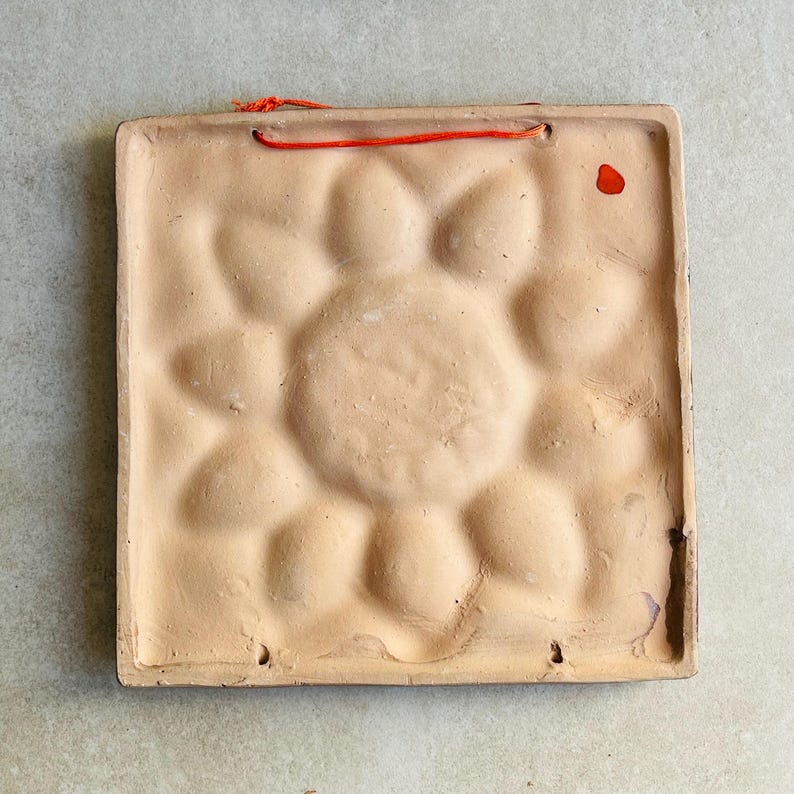 May include: Back of a square, beige ceramic tile with a raised floral design. The tile has a rough texture and a small, red string for hanging. There are two small holes at the bottom and a red paint mark on the right side.