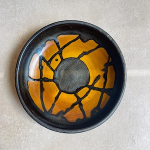 Puede incluir: Un plato redondo de cerámica con un borde negro mate oscuro y una zona circular central. La superficie interior muestra un esmalte amarillo mostaza vibrante contrastado por gruesas líneas negras irregulares que irradian del centro.