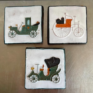 May include: Three vintage ceramic tiles featuring images of antique cars. The tiles are white with black borders and depict a green car, a black and orange car, and a black and orange horse-drawn carriage.