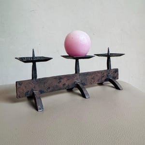 May include: A dark brown, hammered metal candle holder with three square platforms, each featuring a central spike. A smooth, light pink spherical candle rests on the centre platform, while the two outer platforms remain empty.