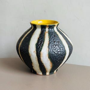 Puede incluir: Un jarrón de cerámica con cuerpo redondeado e interior amarillo. El exterior presenta franjas verticales alternas en blanco y negro, con una superficie texturizada y moteada en las franjas negras. El jarrón es un elemento decorativo para el hogar.