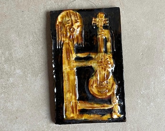 May include: A rectangular ceramic wall hanging featuring a musician playing a stringed instrument. The figure and instrument are in shades of gold and brown against a black background. The piece has a glossy finish and a wire for hanging.