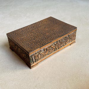 Puede incluir: Una caja rectangular de cobre con tapa texturizada y laterales tallados. La caja tiene un acabado marrón oscuro con un patrón de pequeñas formas irregulares en la tapa. Los laterales presentan tallas en relieve detalladas.