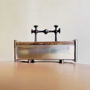 May include: A rectangular metal box with a decorative handle. The box is made of a brushed metal with a brass-coloured top and four short legs. The handle is a dark metal with a cross-like design.