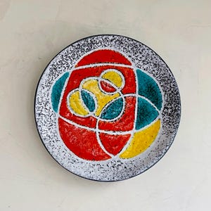 Pode incluir: Um prato redondo decorativo com uma borda preta salpicada e um design abstrato colorido. O design apresenta formas vermelhas, amarelas e turquesas delineadas em branco. O prato tem um acabamento brilhante.