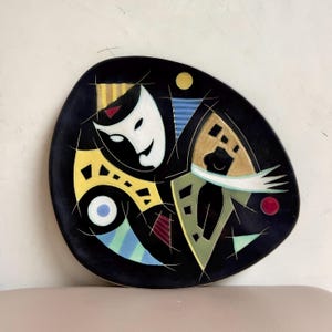 Puede incluir: Un plato decorativo de cerámica negra con un diseño abstracto. El diseño presenta una máscara blanca, formas geométricas amarillas, azules y verdes y detalles en rojo. El plato tiene una forma asimétrica única.