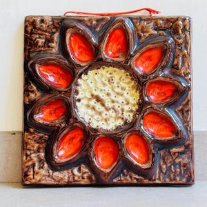May include: Square ceramic tile with a floral design. The tile features a central cream-colored circle surrounded by vibrant orange petal-shaped elements. The background is a textured brown with a glossy finish, and a red string is attached for hanging.