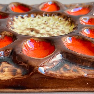 May include: A close-up of a ceramic egg tray with a brown base and vibrant orange and yellow accents. The tray has multiple egg-shaped indentations and a central circular area. The glossy finish reflects light, highlighting the colors and textures.