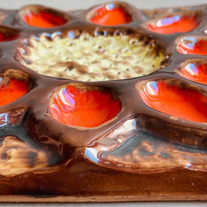 May include: A close-up of a ceramic egg tray with a brown base and vibrant orange and yellow accents. The tray has multiple egg-shaped indentations and a central circular area. The glossy finish reflects light, highlighting the colors and textures.