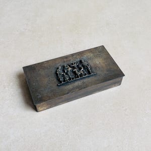 May include: A rectangular, tarnished metal box with a raised relief of a group of people and a dog. The box has a hinged lid and a slightly distressed appearance, suggesting age. The relief is centered on the lid.