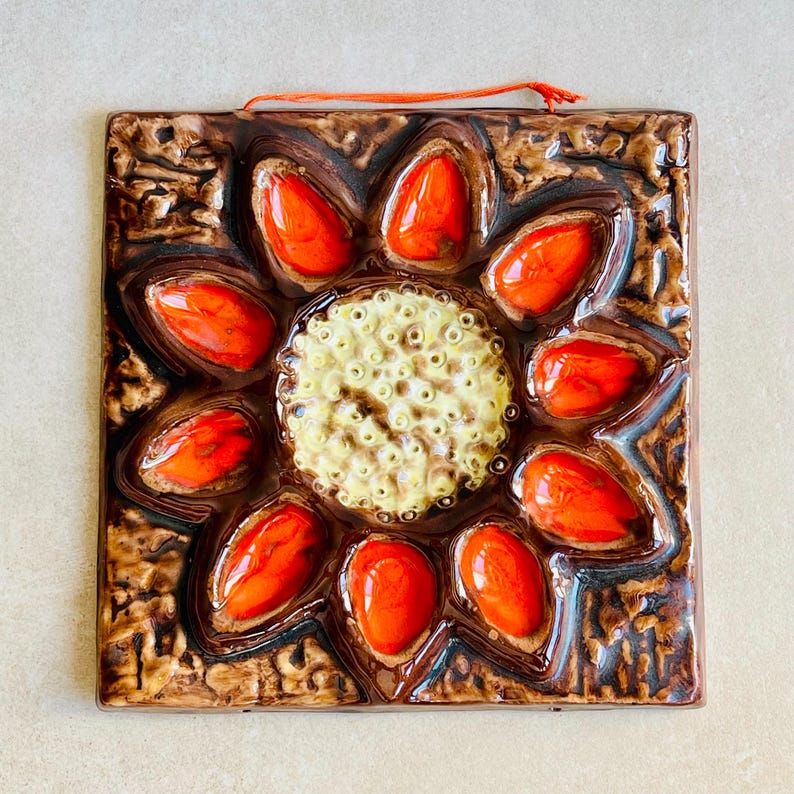 May include: Square ceramic tile with a floral design. The tile features a central cream-colored circle with small holes, surrounded by ten orange petal-shaped elements. The background is brown with a textured pattern, and an orange string is attached for hanging.