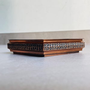 May include: A square, low-profile copper-toned metal tray with a flat top surface and a decorative side band featuring a repeating, dark-toned, textured geometric pattern. The object has a metallic finish with visible highlights and shadows.