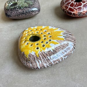 Könnte beinhalten: Eine Sammlung dekorativer Keramik-Blumenfrösche. Der Hauptfrosch ist oval mit einem gelb-weißen Blumenmuster und einem zentralen Loch. Die anderen Frösche haben braune, grüne und rote Craquelé-Muster.