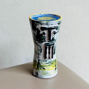 Puede incluir: Un jarrón cilíndrico de cerámica con un diseño de rostro abstracto pintado a mano. El exterior muestra una base blanca con pinceladas superpuestas en verde oscuro y amarillo. El borde superior es amarillo y el interior es de color azul vivo.
