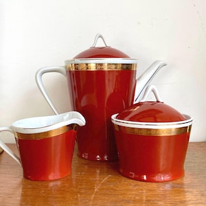 May include: A red and white porcelain tea set with gold trim. The set includes a teapot, creamer, and sugar bowl.