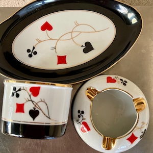 Pode incluir: Um conjunto de cinzeiros de porcelana vintage com acabamento preto e dourado. O conjunto inclui uma bandeja oval grande, um cinzeiro redondo pequeno e um porta-cigarros cilíndrico. Cada peça apresenta um fundo branco com um design preto e dourado de naipes de cartas.