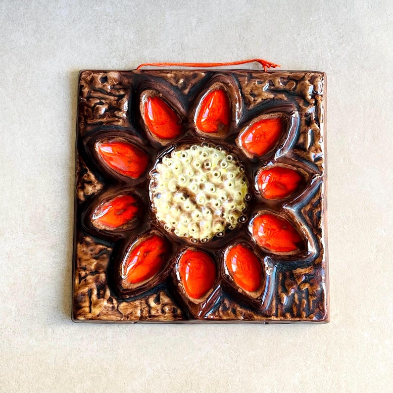 May include: A square, ceramic wall hanging featuring a sunflower design. The flower has bright orange petals surrounding a textured, cream-colored center. The background is a textured brown with an orange string for hanging.