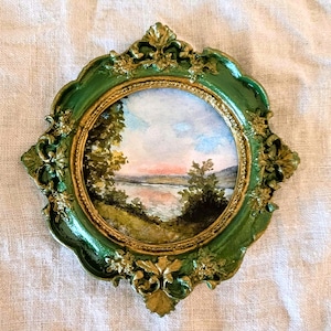 Puede incluir: Una pintura de acuarela de una escena de lago con árboles y un cielo rosa, enmarcada en un marco ovalado verde y dorado.