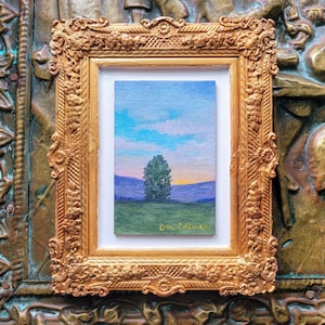 May include: A small landscape painting in a gold ornate frame. The artwork depicts a tree in a green field with purple mountains under a blue sky with clouds. The artist's signature is in the lower right corner.