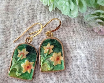 Può includere: Un paio di orecchini pendenti dorati con sfondo verde e narcisi gialli. Gli orecchini sono in metallo e hanno una chiusura a gancio.