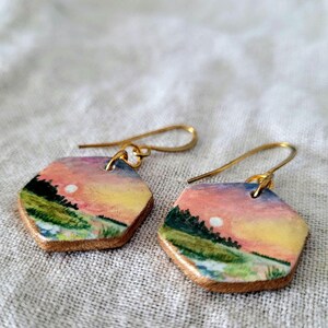 Könnte beinhalten: Ein Paar sechseckige Ohrringe mit einem gemalten Sonnenuntergangs-Motiv. Die Ohrringe sind aus Holz und haben goldfarbene Haken.