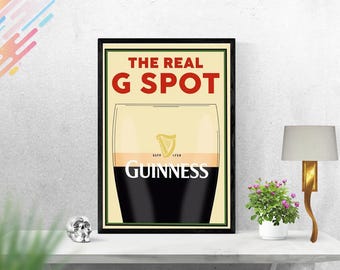 Op de afbeelding: Een ingelijste poster met de tekst "THE REAL G SPOT" in het rood en een afbeelding van een Guinness-pint. Het Guinness-logo is zichtbaar. De poster zit in een zwarte lijst en staat op een witte tafel.