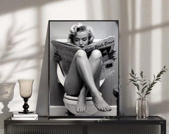 May include: A black and white framed wall art print featuring a person with blonde hair sitting on a toilet while reading a newspaper titled New York Times. The person is positioned with legs crossed, holding the open newspaper in front of their face.