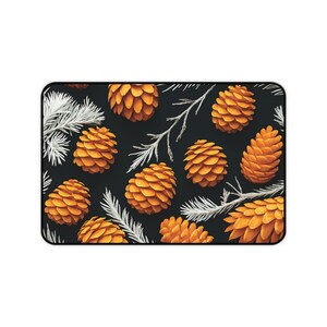 May include: Rectangular mouse mat with a repeating pattern of orange pine cones and white pine needles on a black background. The design is a nature-inspired illustration, suitable for a desk accessory.