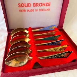May include: A set of five solid bronze spoons with colourful enamelled handles in a red presentation box. The interior lid text reads SOLID BRONZE HAND MADE IN THAILAND. Each spoon features a unique coloured handle with intricate gold detailing.