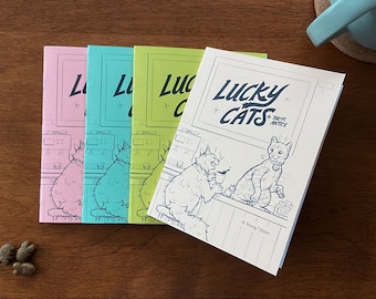 Op de afbeelding: Vier verschillende gekleurde stripboeken op elkaar gestapeld. Het bovenste boek is wit met zwarte tekst die "Lucky Cats" en "A Young Chicken Comic" zegt. De andere boeken zijn roze, blauw en groen met zwarte tekst die "Lucky Cats" zegt.