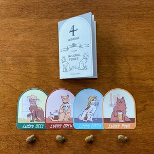 May include: Four illustrated stickers featuring cats with different themes: Lucky Bees, Lucky Brew, Lucky Catch, and Lucky Pour. The stickers are in a rounded arch shape with a light brown background. The stickers are arranged in a row on a wooden surface. A small booklet titled "4 Adventures: Fantastical Felines" is in the background.