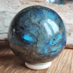 Peut inclure: Une sphère en labradorite polie, révélant des reflets irisés bleus et turquoise sur une surface gris foncé et noire. La boule de pierre précieuse repose sur un petit socle circulaire blanc, soulignant sa forme sphérique.