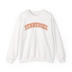 May include: A white long-sleeve crewneck sweatshirt featuring the word TENNESSEE arched across the chest in a block font. The letters are filled with a repeating pattern of small orange and light brown checkered and floral squares.