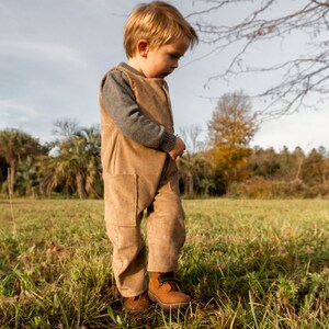 May include: A young child wearing a brown corduroy overall jumpsuit and brown shoes stands in a grassy field. The child is looking down at their feet.