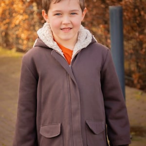 May include: A young person wearing a brown jacket with a fleece lining and a zipper closure. The jacket has two pockets on the front. The person is wearing blue jeans and has short brown hair.