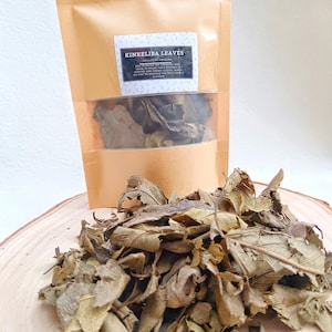 Op de afbeelding: Een bruine papieren verpakking van Kinkeliba-bladeren met een doorzichtig venster en een stapel gedroogde bladeren op een houten oppervlak. De verpakking heeft een label met de tekst "KINKELIBA LEAVES". De bladeren zijn een mix van bruin en groen.