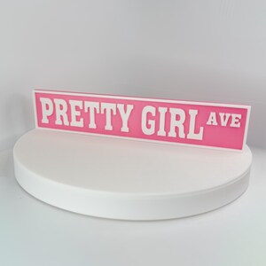 May include: A pink street sign with the words "PRETTY GIRL AVE" in white block letters. The sign is displayed on a white, circular platform against a white background. The sign is a decorative item.