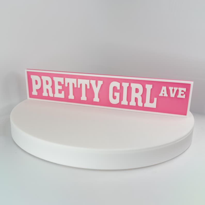 May include: A pink street sign with the words "PRETTY GIRL AVE" in white block letters. The sign is displayed on a white, circular platform against a white background. The sign is a decorative item.