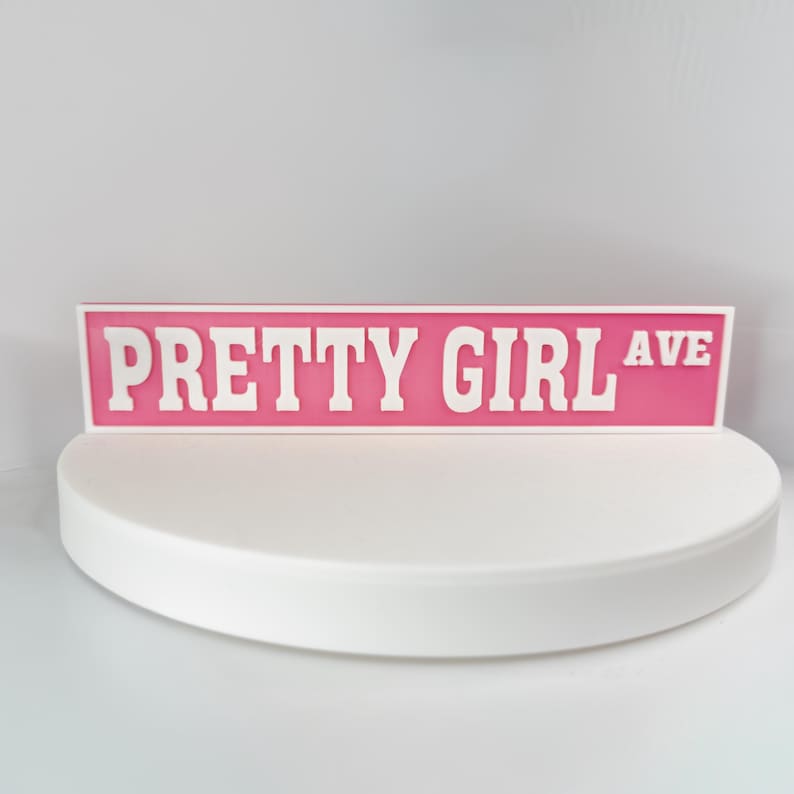 May include: A pink street sign with the words "PRETTY GIRL AVE" in white block letters. The sign is rectangular and set against a white background. The sign is displayed on a white, semi-circular platform.