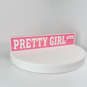 May include: A pink street sign with the words "PRETTY GIRL AVE" in white block letters. The sign is displayed on a white, circular platform against a white background. The sign is a decorative item.