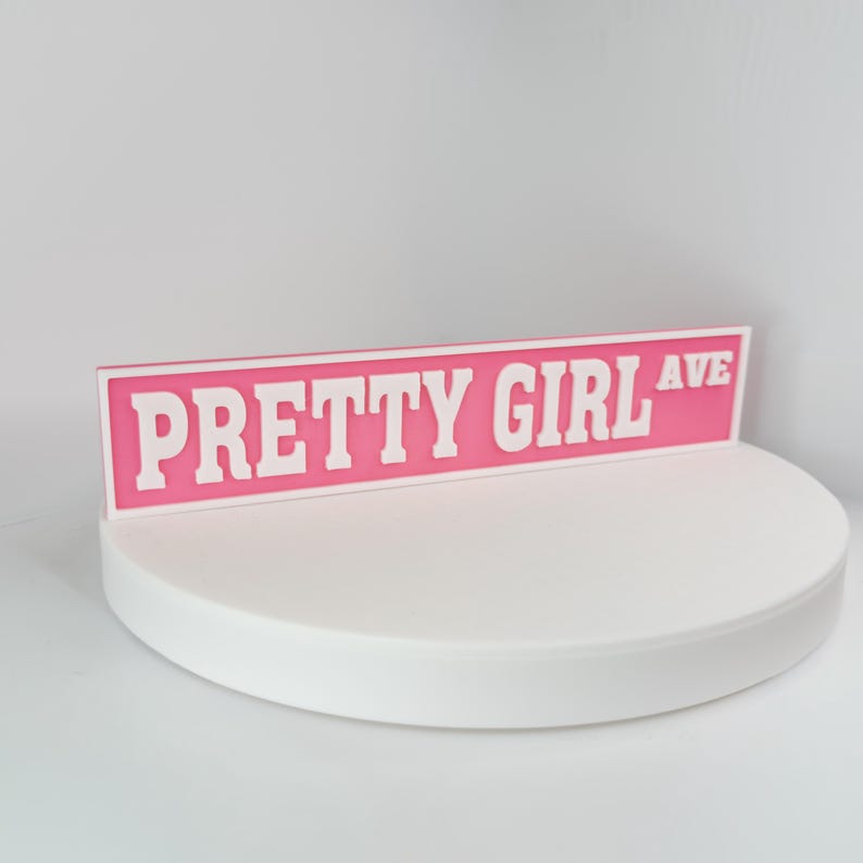 May include: A pink street sign with the words "PRETTY GIRL AVE" in white block letters. The sign is displayed on a white, circular platform against a white background. The sign is a decorative item.