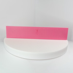 May include: A rectangular, bright pink wall-mounted shelf with a keyhole for hanging. The shelf is displayed on a white, circular platform against a white backdrop. The shelf is a solid color and has a smooth surface.