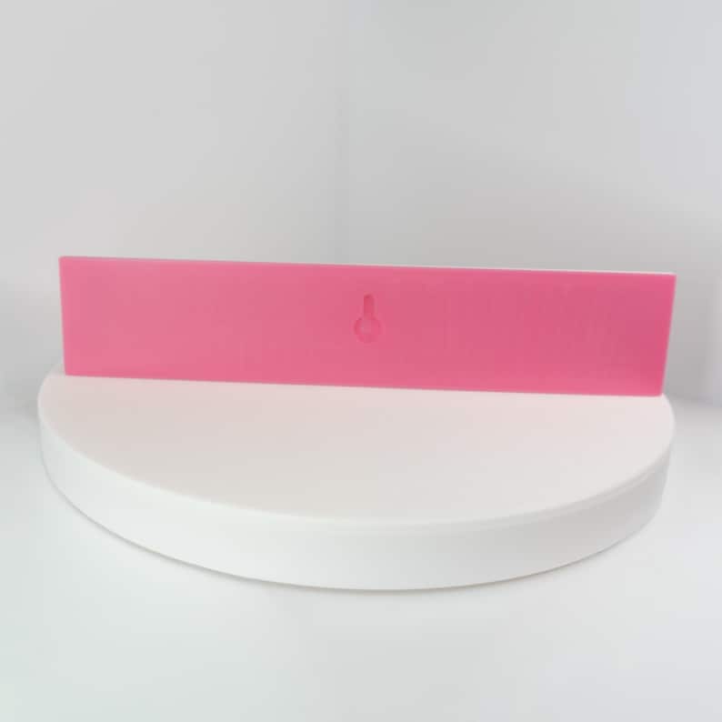May include: A rectangular, bright pink wall-mounted shelf with a keyhole for hanging. The shelf is displayed on a white, circular platform against a white backdrop. The shelf is a solid color and has a smooth surface.