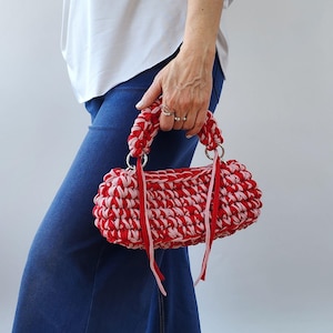 Könnte beinhalten: Eine rot-rosa gehäkelte Handtasche mit silbernen Metallringen und Griffen. Die Tasche wird von einer Person gehalten, die eine blaue Jeans und ein weißes Hemd trägt. Die Tasche hat eine zylindrische Form und besteht aus einem gewebten Material.